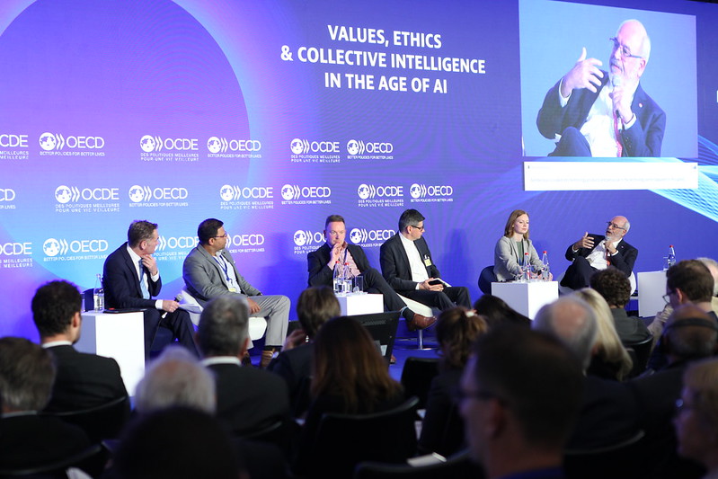 A photo of a group of people smartly dressed sitting as a panel on a stage. There is a large blue background behind them with text that reads 'Values, Ethics, and Collective Intelligence in the Age of AI', along with the OECD logo. There is an audience watching them speak.