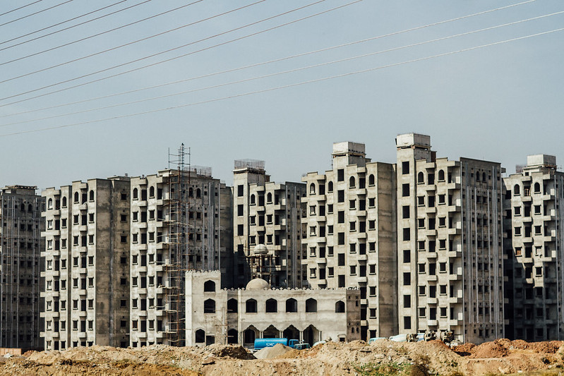 A photo of a line of grey apartment buildings which are under construction. The sky is blue and the ground is rocky and brown. There are cables in the foreground.