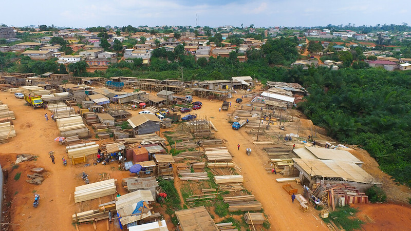A photo of dozens of small cabins with metal roofs containing large logs of wood. They are sitting on a beige coloured hill. There are trees dotted around and a collection of houses behind the cabins.
