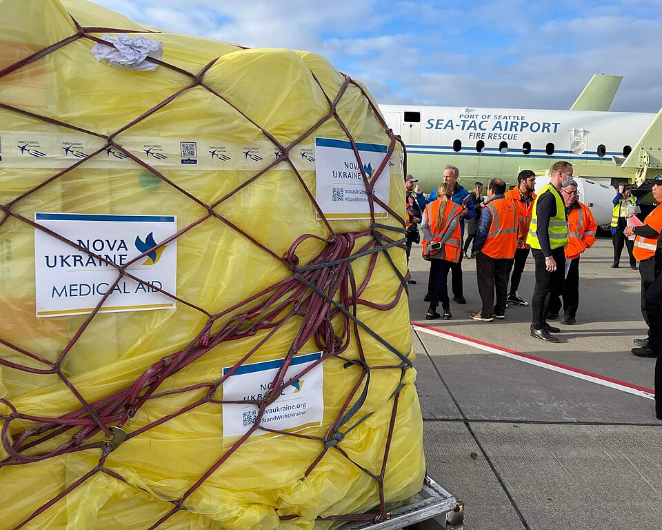 A photo of medical aid being sent to Ukraine from Seattle, United States. There is a very large aid box wrapped in a yellow sheet and covered by a net in the foreground of the photo. In the background there is an aeroplane with some people in hi-vis orange vests standing next to it.