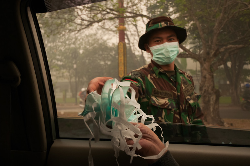 A photo from inside of a car of a person in an army uniform standing outside of a car and handing some blue medical masks to someone inside the car. There is smoke all around them with some trees.