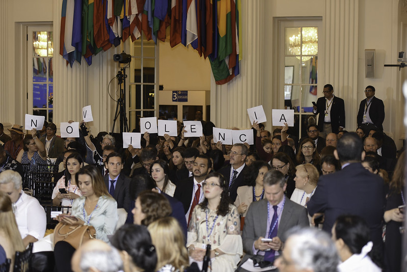 A photo of a group of people sitting in chairs in a conference room, facing towards the camera. Behind them, people hold up signs that read 'No Violencia' in Spanish. There are some flags drooping behind them.