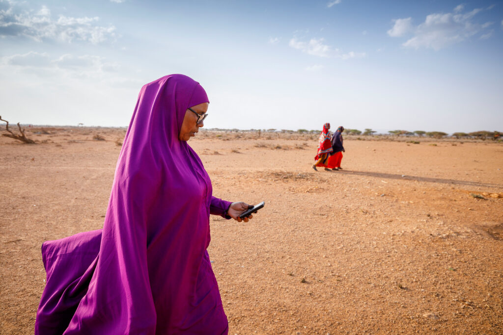 A photo of three people walking through a desert landscape. The person in the foreground wears purple and looks at the mobile phone in their hand, and the two people in the background wear red. 