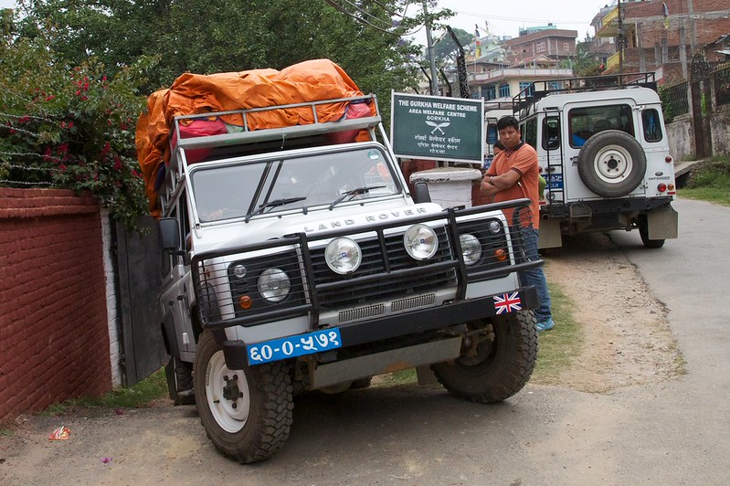 A photo of a 4x4 vehicle making a sharp turn on a narrow road. The vehicle is grey and it has orange sacks on the roof. A man stands next to the car looking at the camera. There is another 4x4 in the background.