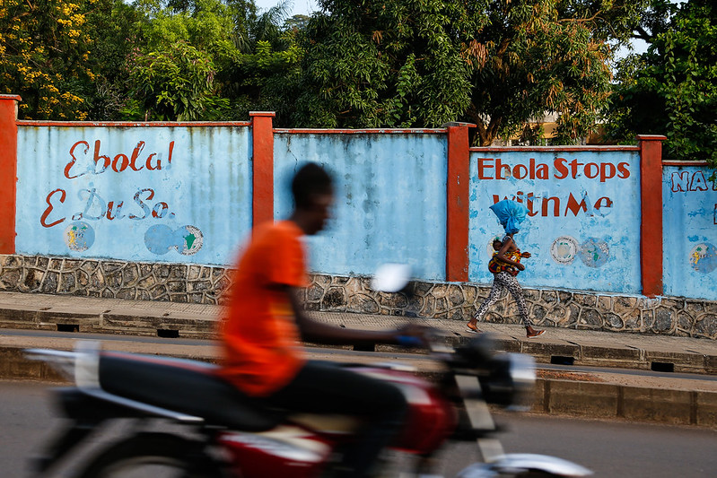 A photo of a mural on a wall next to a road. The mural has a blue background with red posts, and red text that reads 'Ebola! E Du So. Ebola Stops With Me'. People walk past and a man on a motorbike zooms past.