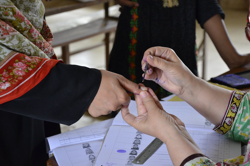 A photo of a person holding another person's hand, marking the index finger with black ink to indicate they voted. Underneath their hands are voting slips.
